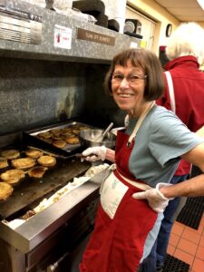 image of an older woman wearing a red apron, and standing at a large stove cooking food.